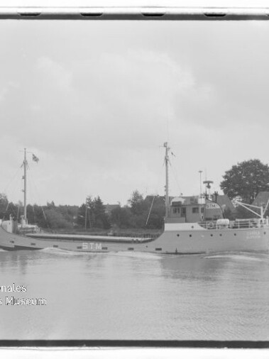 Schwarzweißaufnahme eines kleinen Frachtschiffs mit Schriftzug STN auf ruhigem Fluss, Bäume und Häuser im Hintergrund; Wasserzeichen des Internationalen Maritimen Museums Hamburg.