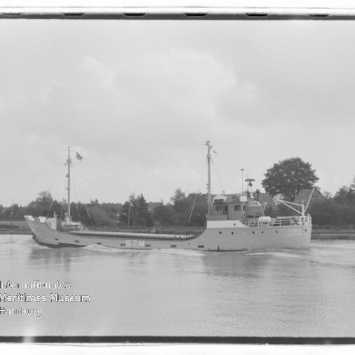 Schwarzweißaufnahme eines kleinen Frachtschiffs mit Schriftzug STN auf ruhigem Fluss, Bäume und Häuser im Hintergrund; Wasserzeichen des Internationalen Maritimen Museums Hamburg.