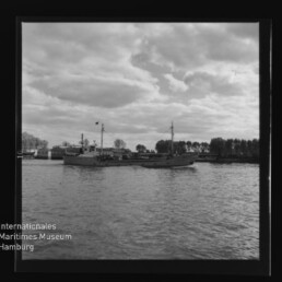 Frachtschiff auf ruhigem Hafenwasser vor bewölktem Himmel; links unten Logo und Schriftzug des Int. Maritimen Museums Hamburg