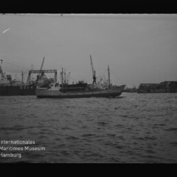 Kleiner Frachter im Hafen vor Kränen und Dockanlagen; unten links das Logo des Internationalen Maritimen Museums Hamburg.