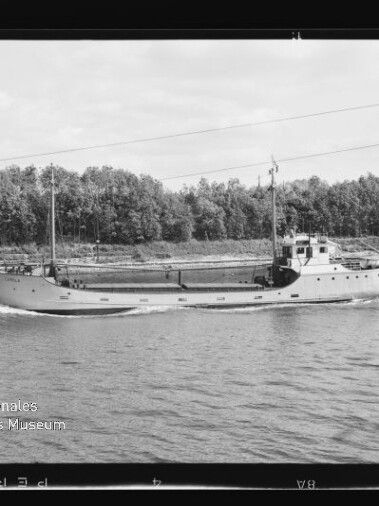 Schwarzweißfoto eines kleinen Frachtschiffs, fahrend auf einem Kanal, Baumreihe am Ufer und Museumsemblem unten links