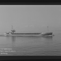 Schwarzweiß-Foto eines Frachtschiffs in Seitenansicht auf ruhigem Meer; Filmrand und Museumswasserzeichen sichtbar.