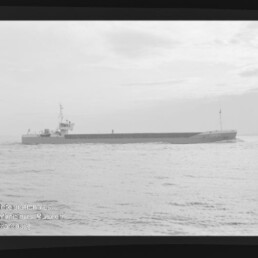 Langgestrecktes Frachtschiff mit niedrigem Aufbau auf ruhiger See unter hellem Himmel, schwarzweißes Foto.