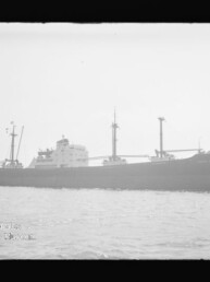 Schwarzweißaufnahme eines Frachtschiffs mit Aufbauten und Ladegeschirr auf ruhigem Wasser.