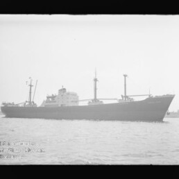 Schwarzweißaufnahme eines Frachtschiffs mit Aufbauten und Ladegeschirr auf ruhigem Wasser.