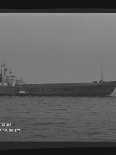 Großes Frachtschiff mit Schriftzug CARRIER auf ruhiger See, kleines Begleitboot links und Museumslogo unten links