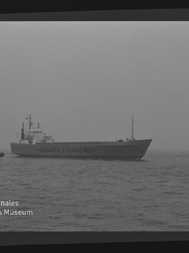 Frachtschiff mit Aufschrift HOLMEN CARRIER und kleinem Schlepper auf ruhiger, nebliger See.