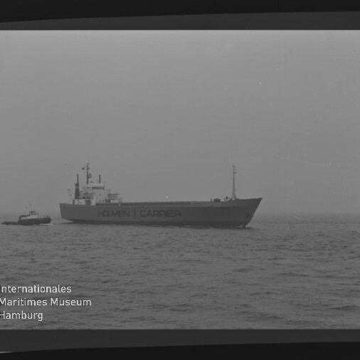 Frachtschiff mit Aufschrift HOLMEN CARRIER und kleinem Schlepper auf ruhiger, nebliger See.