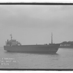 Großes Frachtschiff auf ruhigem Wasser vor bewaldeter Küste; unten links Stempel Internationales Maritimes Museum Hamburg.