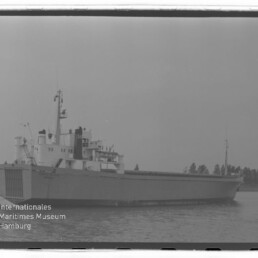 Frachtschiff mit vorderer Rampe und Aufbauten, ruhig auf dem Wasser vor einer Baumreihe; Wasserzeichen Maritimes Museum Hamburg.