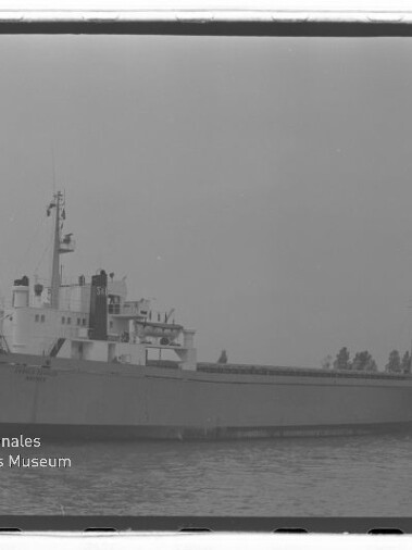 Frachtschiff mit vorderer Rampe und Aufbauten, ruhig auf dem Wasser vor einer Baumreihe; Wasserzeichen Maritimes Museum Hamburg.