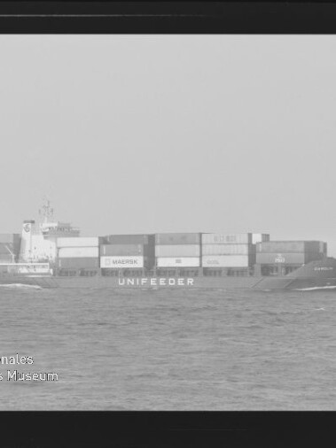 Schwarzweißfoto eines Containerschiffs mit Schriftzug UNIFEEDER und gestapelten Containern auf ruhiger See, Wasserzeichen links unten.