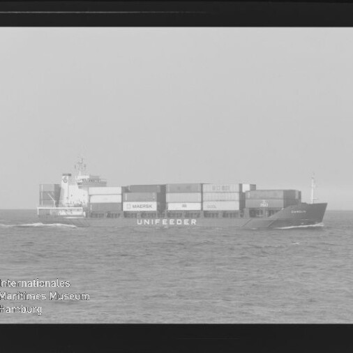Schwarzweißfoto eines Containerschiffs mit Schriftzug UNIFEEDER und gestapelten Containern auf ruhiger See, Wasserzeichen links unten.