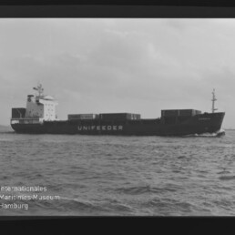 Containerschiff mit Schriftzug UNIFEEDER fährt über ruhiges Meer unter bewölktem Himmel; Museumswasserzeichen unten links