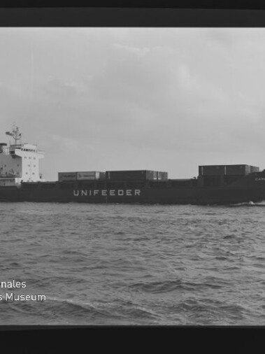 Containerschiff mit Schriftzug UNIFEEDER fährt über ruhiges Meer unter bewölktem Himmel; Museumswasserzeichen unten links