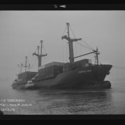 Frachtschiff CAROLINA mit Containern und Ladekränen, begleitet vom Schlepper, auf nebligem Wasser; Museumslogo unten links.