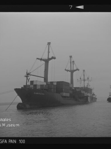 Nebliger Hafen mit einem beladenen Frachtschiff, sichtbaren Containern und Kränen, begleitet von einem Schlepper.