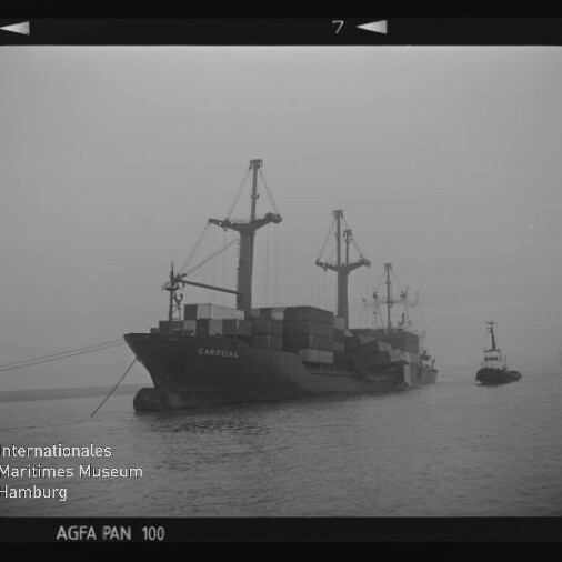 Nebliger Hafen mit einem beladenen Frachtschiff, sichtbaren Containern und Kränen, begleitet von einem Schlepper.