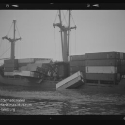 Frachtschiff mit umgestürzten Containern, mehrere Container ins Wasser gefallen, Kräne und gestapelte Container im Hintergrund.