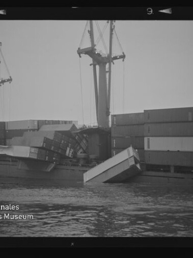 Frachtschiff mit umgestürzten Containern, mehrere Container ins Wasser gefallen, Kräne und gestapelte Container im Hintergrund.