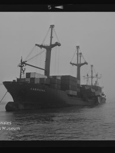 Containerschiff Carolina mit gestapelten Containern und Ladekränen fährt in nebligem Hafen, Museumsemblem unten links.