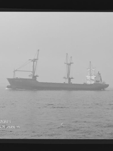 Frachtschiff mit Ladekränen auf ruhiger See bei Dunst, monochrome Aufnahme mit Filmrand und Museumslogo.