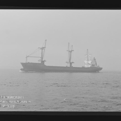 Frachtschiff mit Ladekränen auf ruhiger See bei Dunst, monochrome Aufnahme mit Filmrand und Museumslogo.