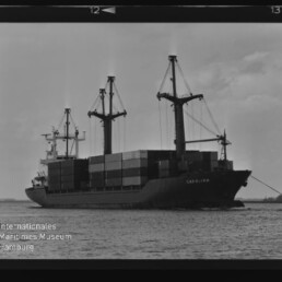 Containerschiff CAROLINA mit gestapelten Containern und Ladekränen auf ruhigem Hafenwasser im schwarzweiß-Filmrahmen.