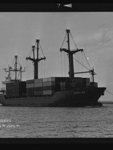 Schwarzweißaufnahme eines Containerschiffs CAROLINA mit drei Ladekränen und gestapelten Containern am Hafen.