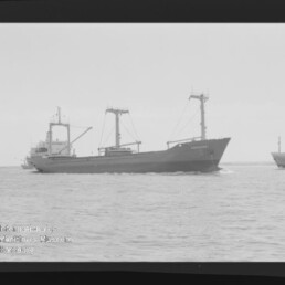 Schwarzweißaufnahme eines Frachtschiffs mit Ladebäumen auf ruhiger See, mit Filmrand (Kodak) und Museumswasserzeichen.