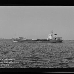 Frachtschiff mit gestapelten Containern quer zur Kamera auf ruhiger See vor flachem Horizont.