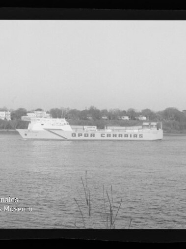 Große Fähre mit auffälligem Schriftzug fährt auf einem Fluss, im Hintergrund Ufer mit Häusern.