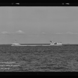 Schwarzweißaufnahme eines langen Frachtschiffs auf ruhiger See am Horizont mit niedrigen Wolken und Wasserzeichen Internationales Maritimes Museum Hamburg.