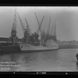 Schwarzweißaufnahme eines Frachtschiffs am Kai vor einer Reihe hoher Hafenkräne, mit kleinem Boot rechts und Museumslogo links unten.