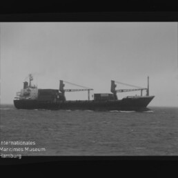 Frachtschiff mit zwei Deckskränen auf See, Horizont und Wasserzeichen des Internationalen Maritimen Museums Hamburg.