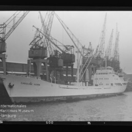 Frachtschiff CAROLINE HORN liegt am Industriequai vor einer Reihe großer Hafenkräne, schwarzweiß.