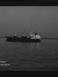 Schwarzweiß-Negativ eines Frachtschiffs mit Ladekränen vor ruhigem Meer und flachem Horizont, Filmrand und Museumslogo sichtbar.