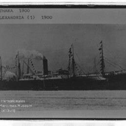 Dampfschiff mit Schornstein und beflaggten Masten; oben sichtbarer Text: ITHAKA 1900, schwarz-weiß.