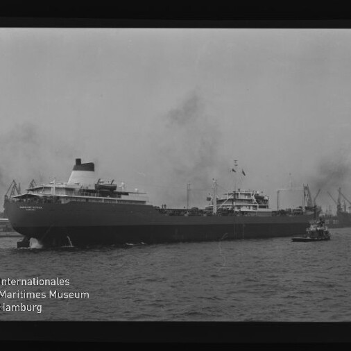 Großes Frachtschiff im Hafen, begleitet von einem Schlepper, mit Hafenkränen im Hintergrund in einer Schwarzweißaufnahme.