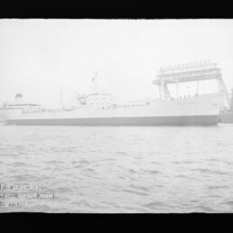 Großes Frachtschiff im Hafen vor Werfthallen mit Kränen, Wellen im Vordergrund und Museumssignet unten links.