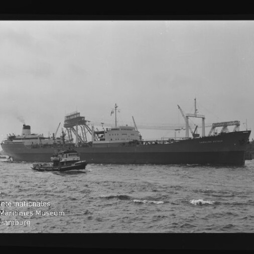 Schwarzweißaufnahme zweier Frachtschiffe im Hafen, im Vordergrund ein Schlepper und im Hintergrund Ladekräne an der Pier.