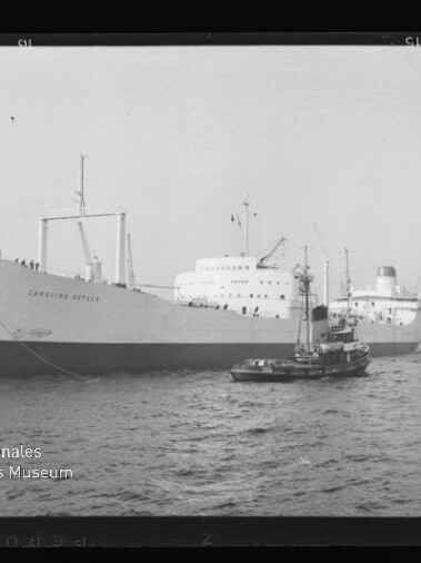 Großes weißes Frachtschiff mit Schlepper im Vordergrund, Hafenkräne im Hintergrund in einer Schwarzweißaufnahme.