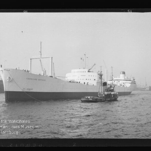 Großes weißes Frachtschiff mit Schlepper im Vordergrund, Hafenkräne im Hintergrund in einer Schwarzweißaufnahme.