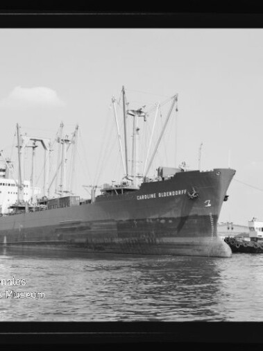 Großes Frachtschiff mit Ladekränen und Schlepper am Bug im Hafen, Schwarzweißaufnahme mit lesbarem Schiffsnamen.