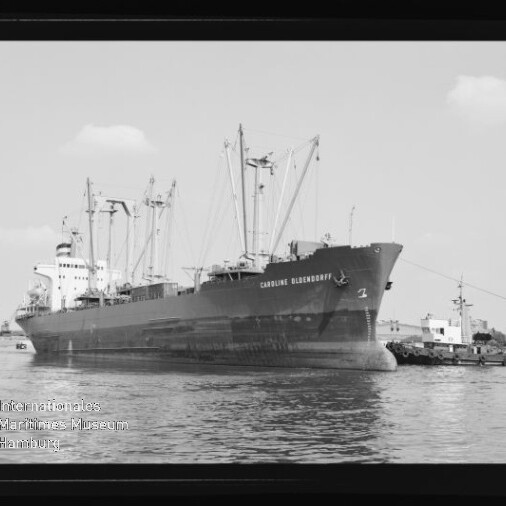 Großes Frachtschiff mit Ladekränen und Schlepper am Bug im Hafen, Schwarzweißaufnahme mit lesbarem Schiffsnamen.