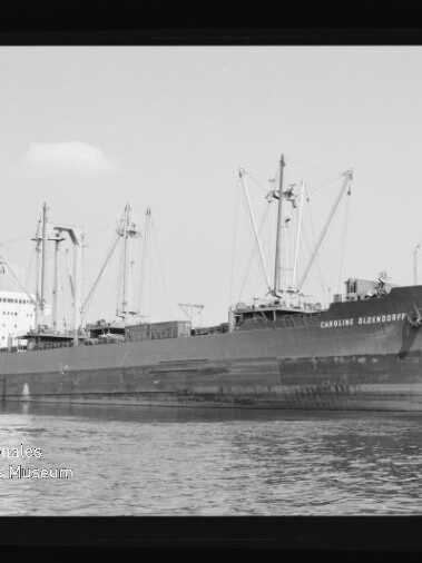 Großes Frachtschiff mit mehreren Ladekränen und gut sichtbarem Schiffsname am Bug, auf ruhigem Wasser vor einem Hafen