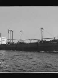 Schwarzweißaufnahme eines Frachtschiffs mit mehreren Ladekränen auf offener See in Seitenansicht, Museumslogo unten links.