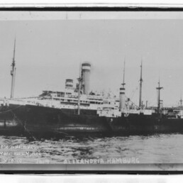 Großes Dampfschiff mit zwei Schornsteinen und mehreren Masten im Hafen; Schriftzug ALEXANDRIA HAMBURG am unteren Bildrand.