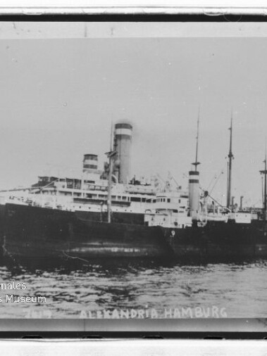 Großes Dampfschiff mit zwei Schornsteinen und mehreren Masten im Hafen; Schriftzug ALEXANDRIA HAMBURG am unteren Bildrand.