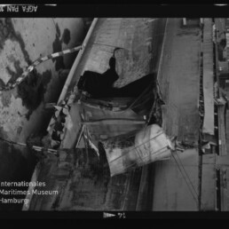 Aufgerissener, verformter Schiffsrumpf mit zerfetzter Metallbeplankung und sichtbaren Tauen, Wasserzeichen 
